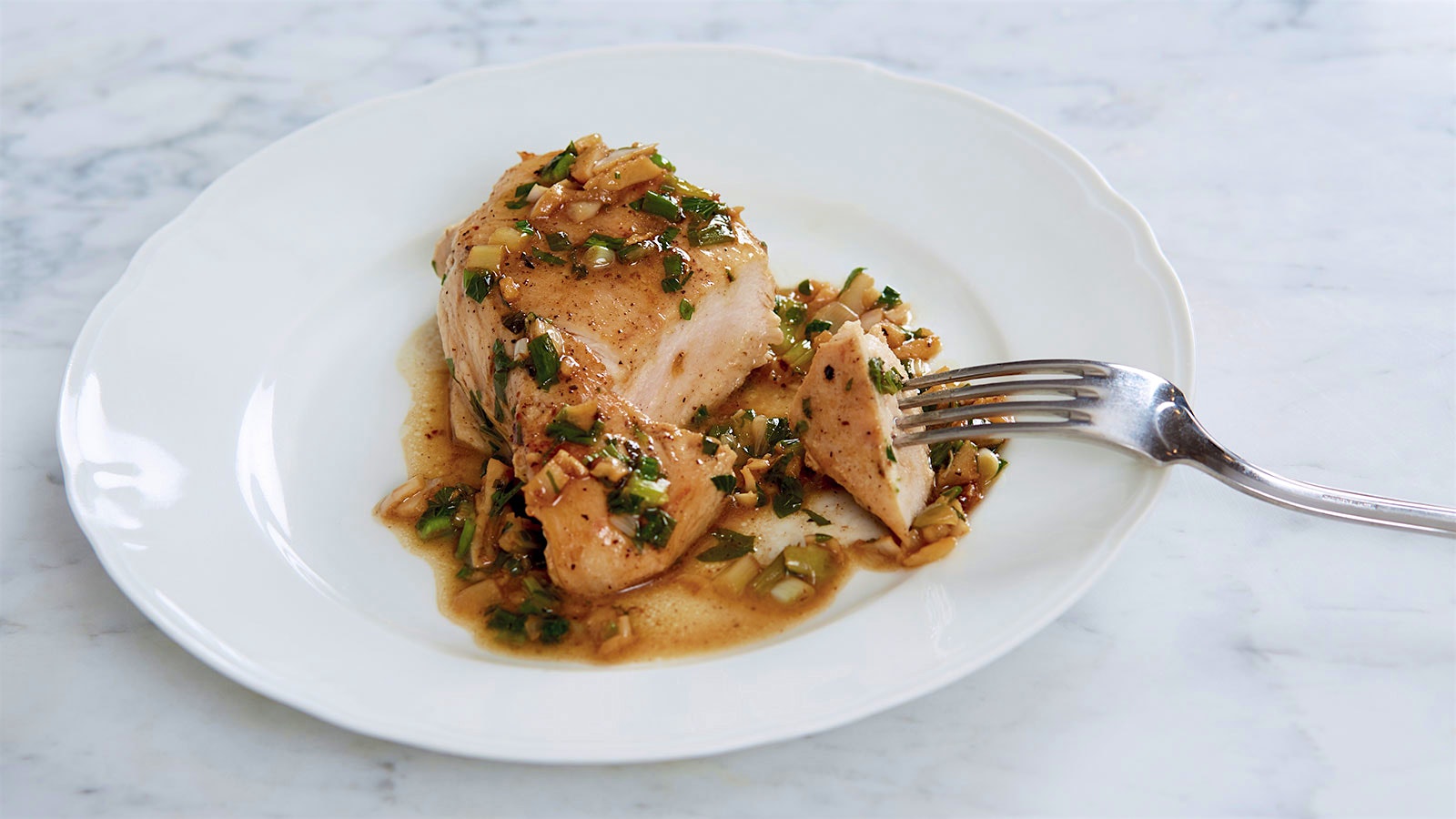 A white plate with a boneless chicken breast topped with an oil-based sauce of garlic, parsley and scallions. 
